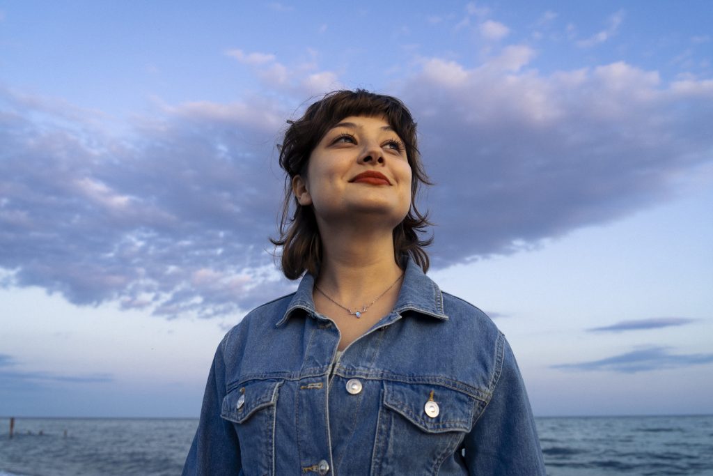 Photograph of a beautiful young woman wearing a denim jacket and smiling towards the sun, taken in the sweet pastel colors of the sunset.